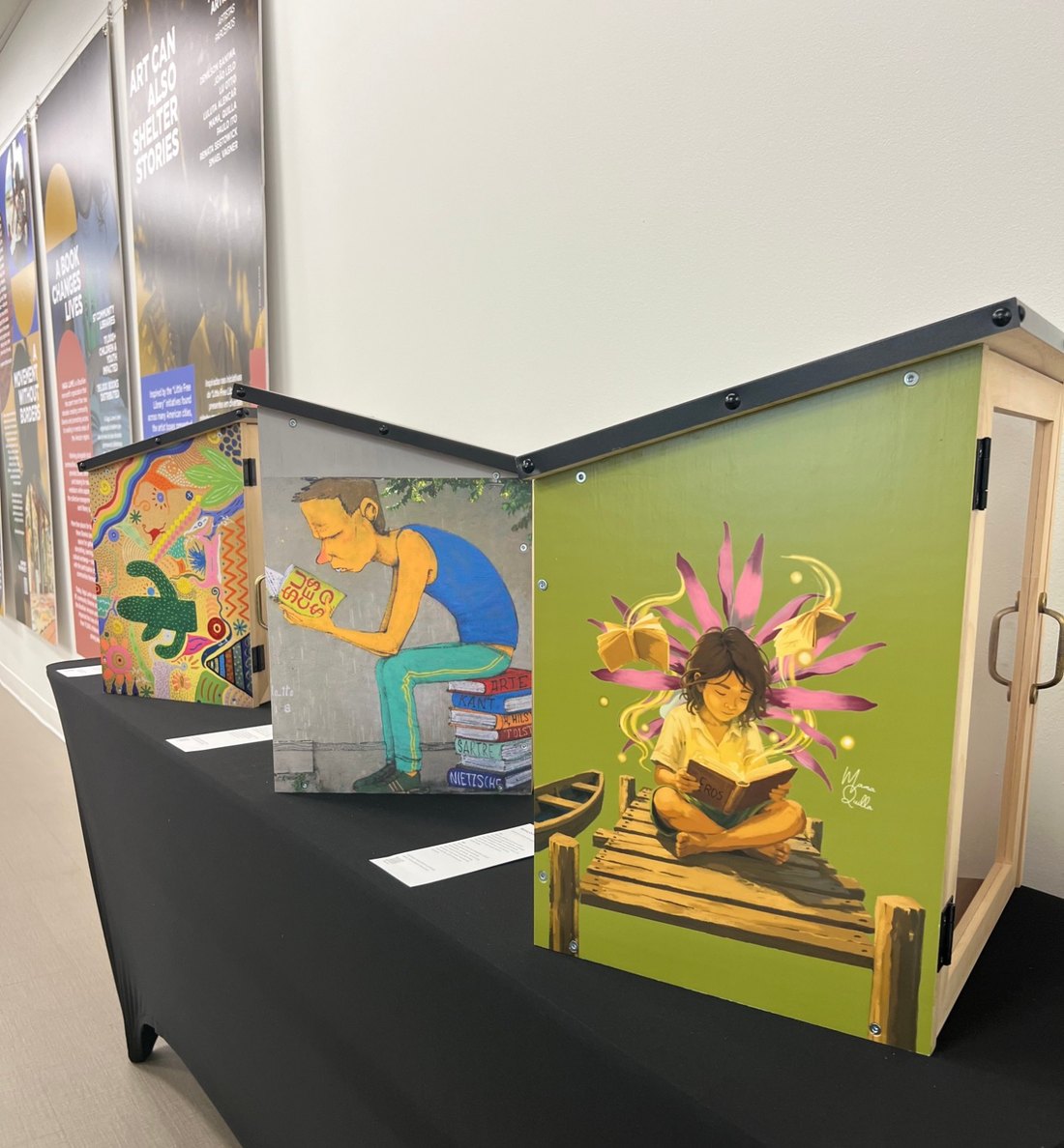 Three painted library boxes on a plinth, with the exhibition's banners on the wall behind them